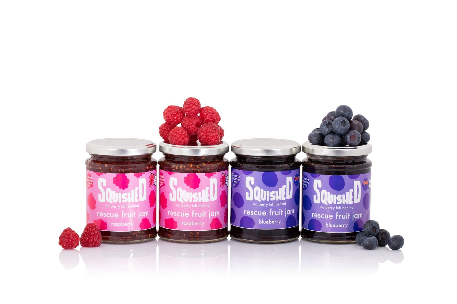 Four jars of fruit preserves with fresh raspberries and blueberries displayed alongside from a product photoshoot.