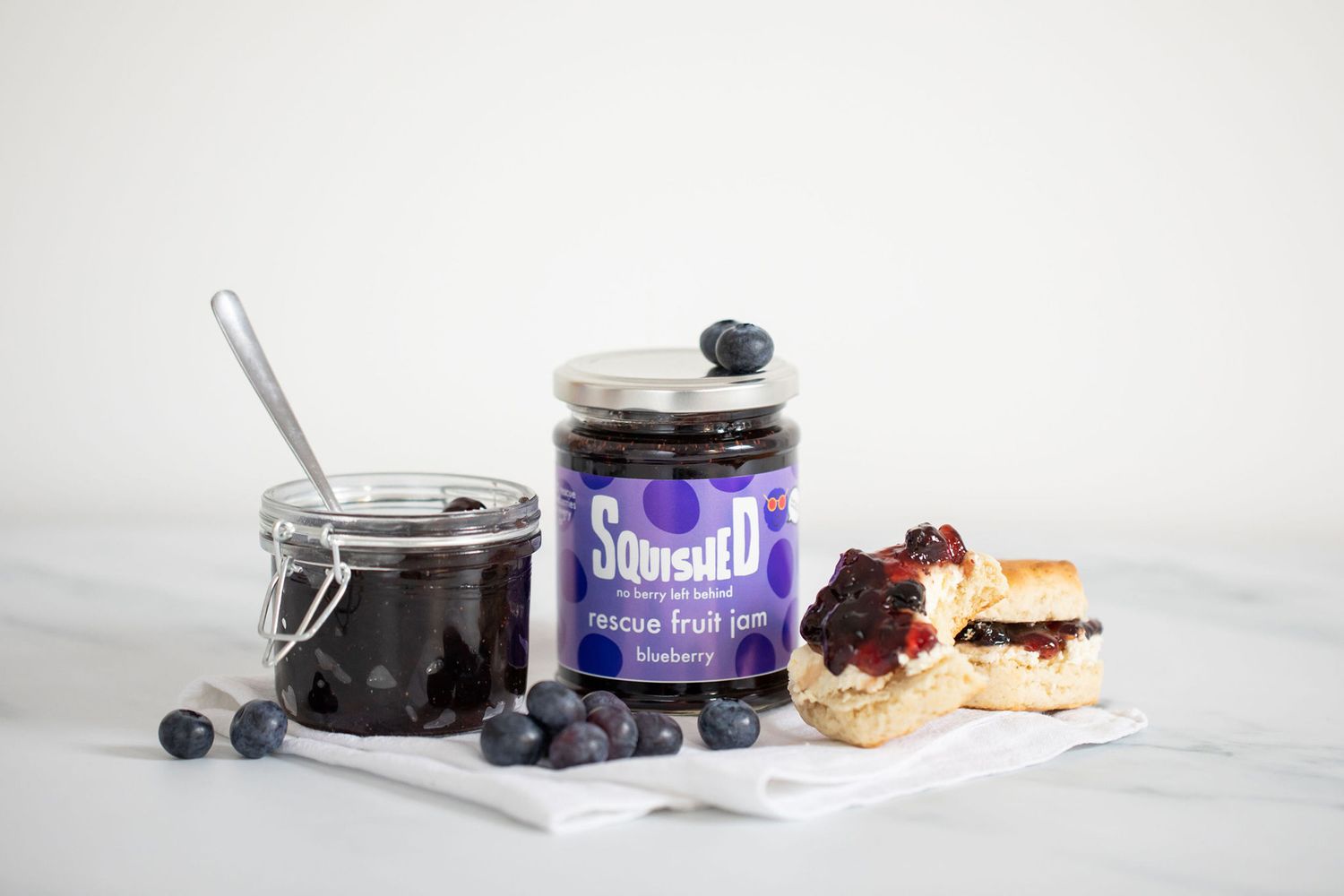 Blueberry preserves in glass jars with fresh berries and pastry on marble counter from a product photoshoot.