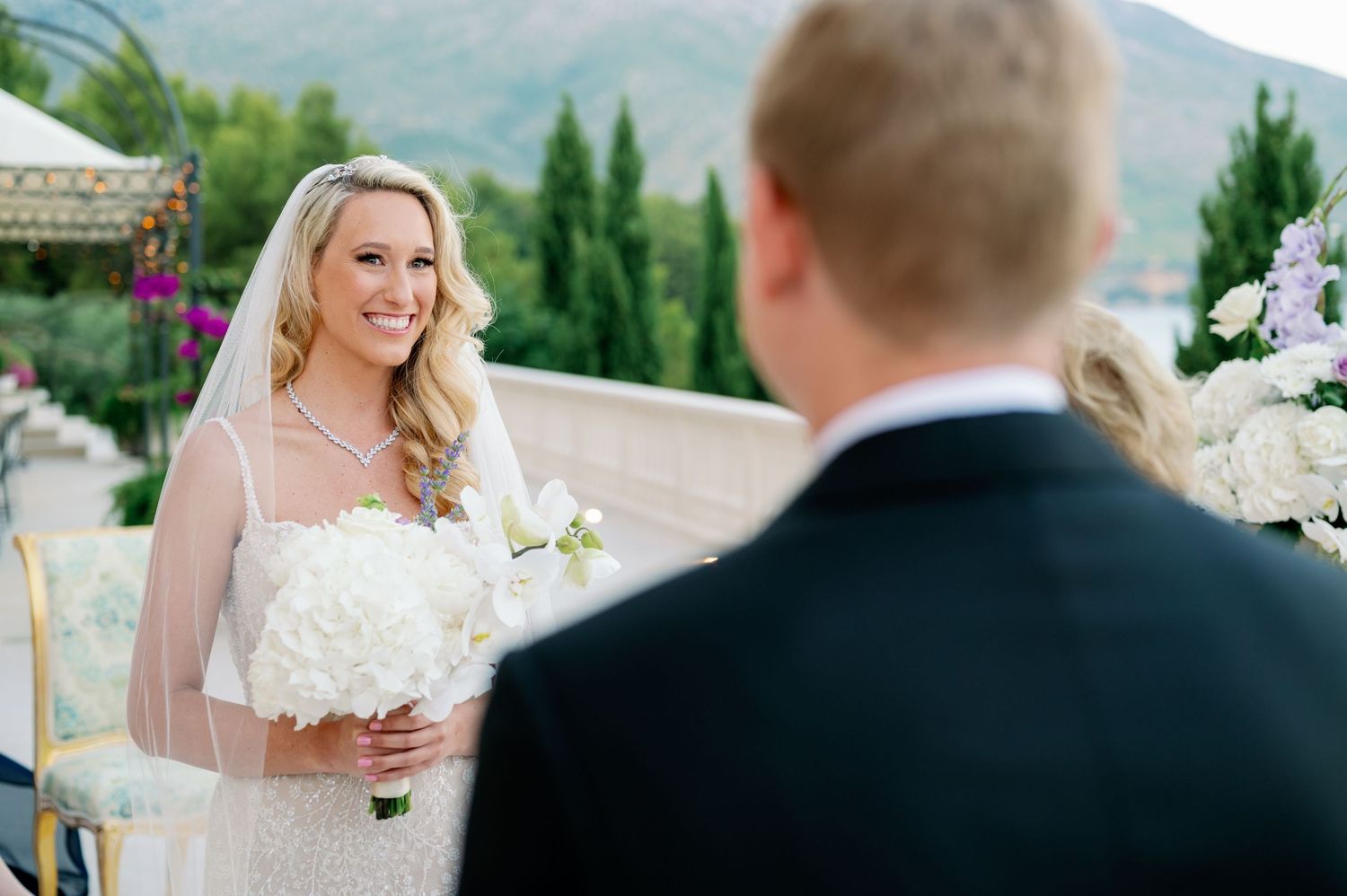 Bride in white lace dress holds bouquet during outdoor ceremony with mountain and cypress tree backdrop.