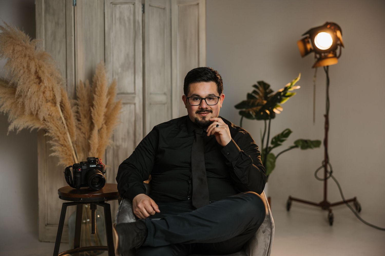 A person in dark clothing sits thoughtfully in a modern room with pampas grass decor and studio lighting.