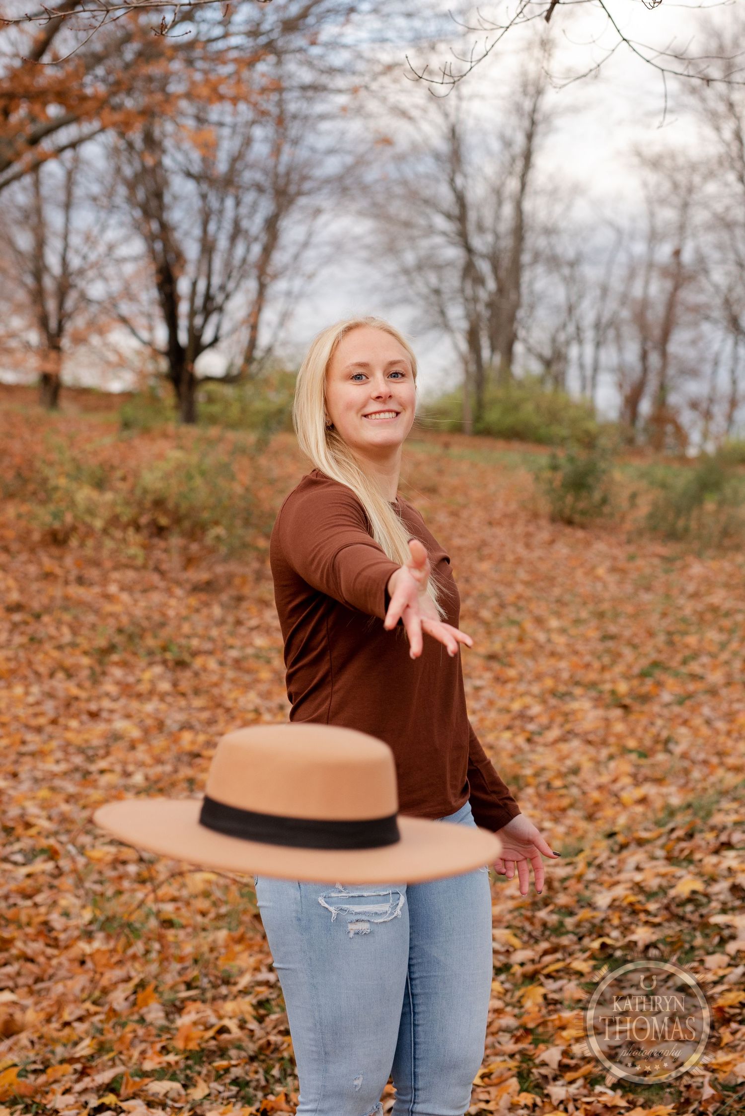 Playful autumn portrait with brown sweater and tossed hat among fall leaves.