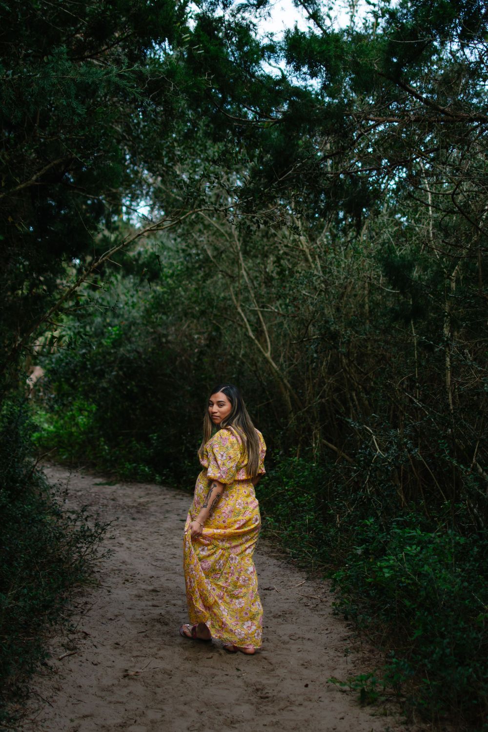 Figure in flowing yellow dress stands on nature path surrounded by lush greenery.