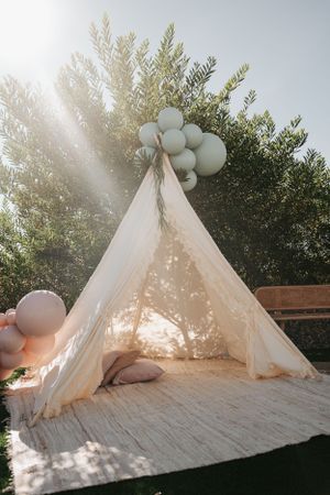 White teepee tent decorated with balloons and plush pillows for baby shower.