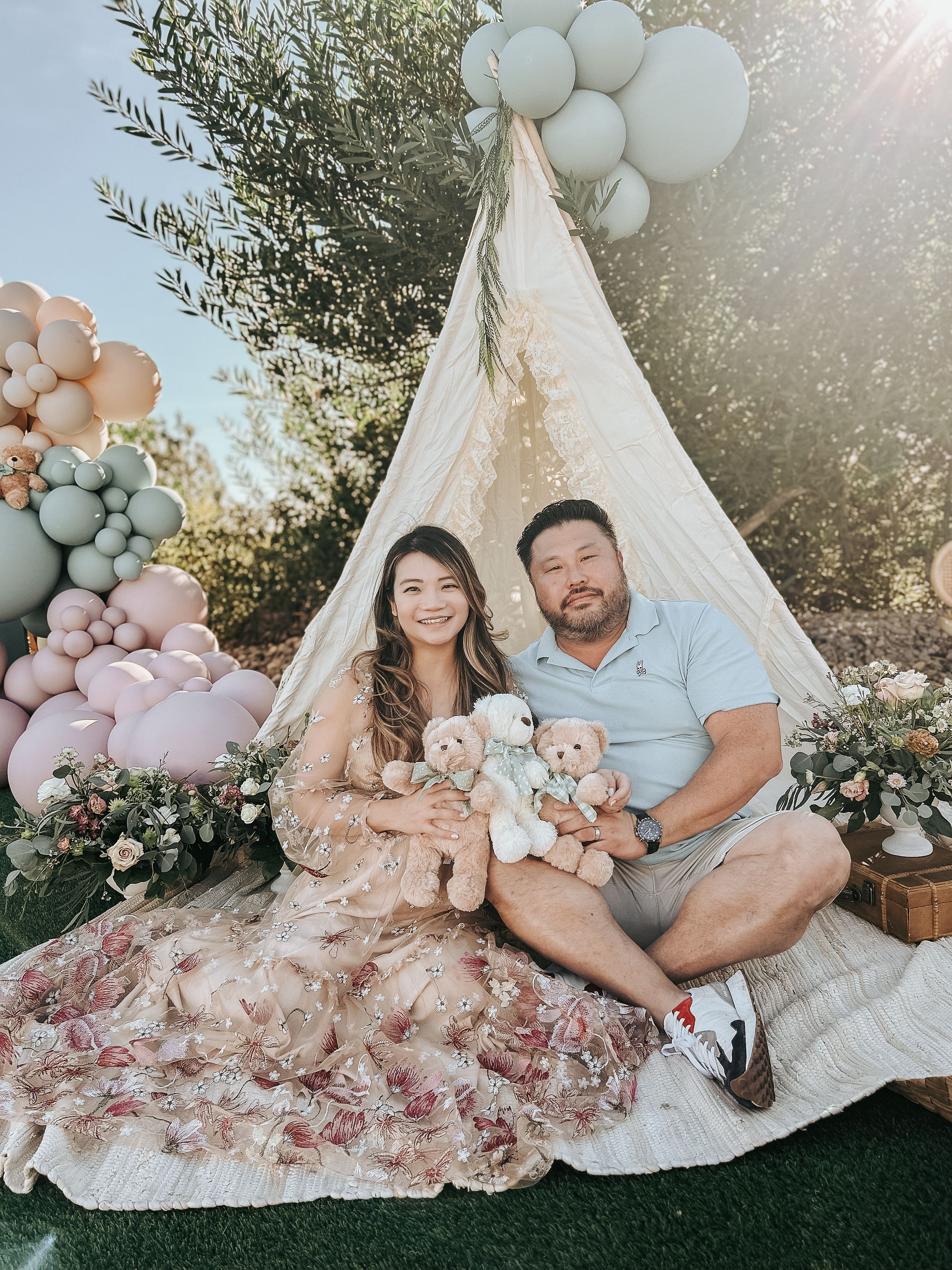 Couple sits together with teddy bears in front of a decorated white teepee with balloons.
