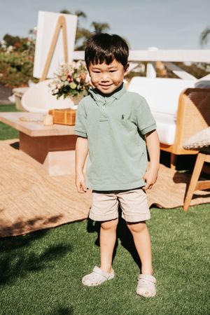 Child in green polo shirt and shorts playing on grass near party setup.