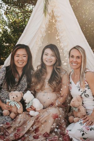 Three friends sitting with teddy bears in front of a teepee at a baby shower.