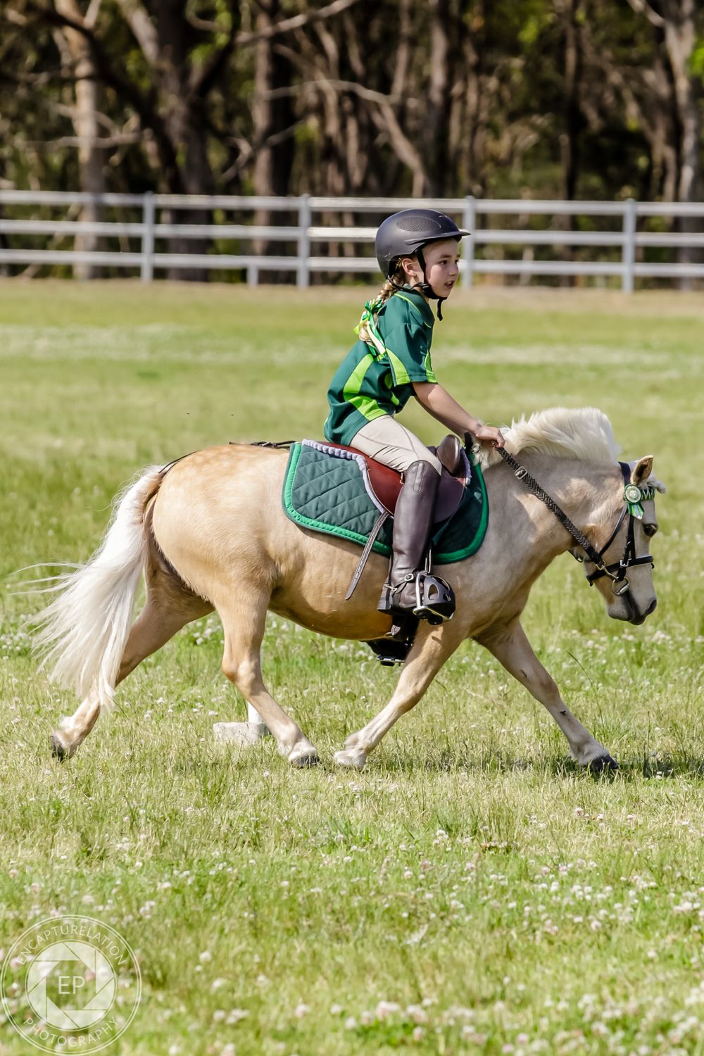 A rider on a palomino pony trots through a grassy field with white fencing in the background.