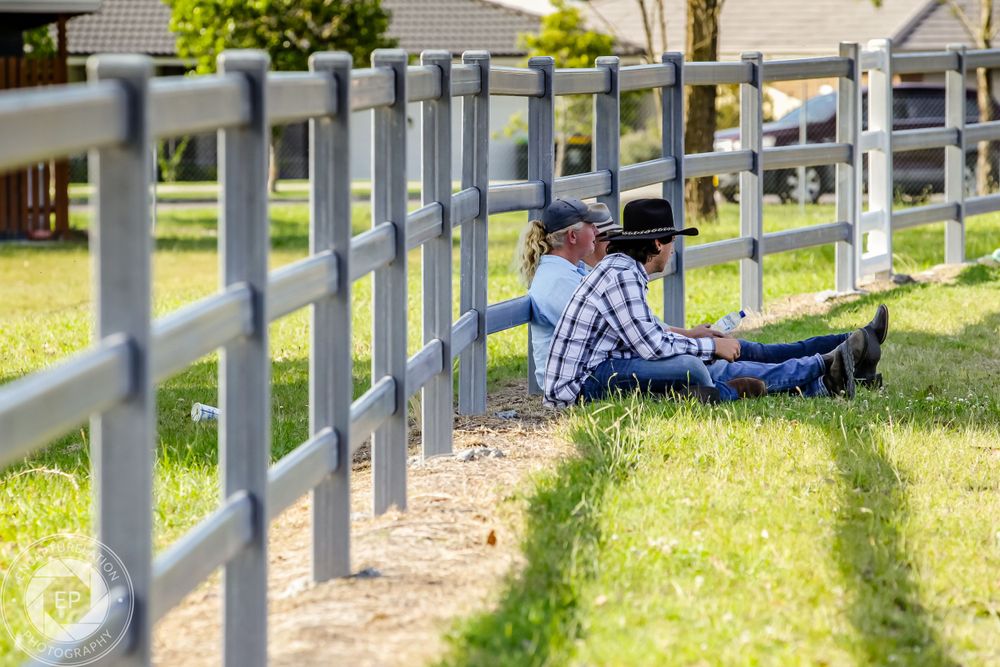 A person rests against a white ranch fence on sunny grass lawn.