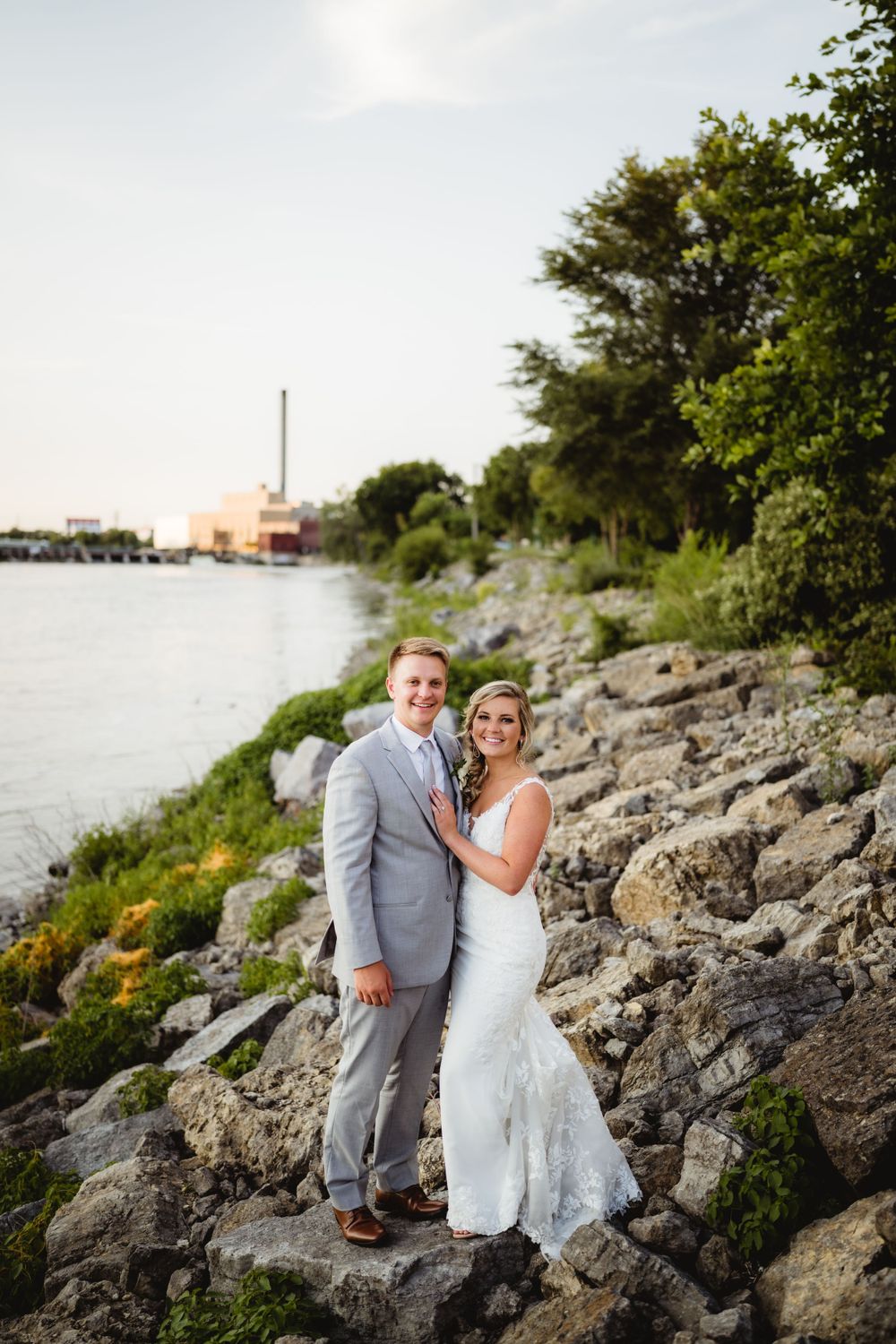 Couple in wedding attire poses together on rocky shoreline with water and buildings in background.