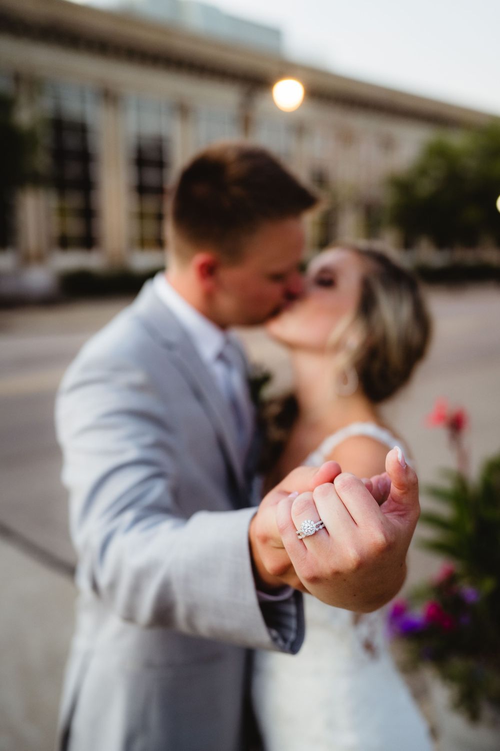 Ring detail shot with couple embracing against urban backdrop.