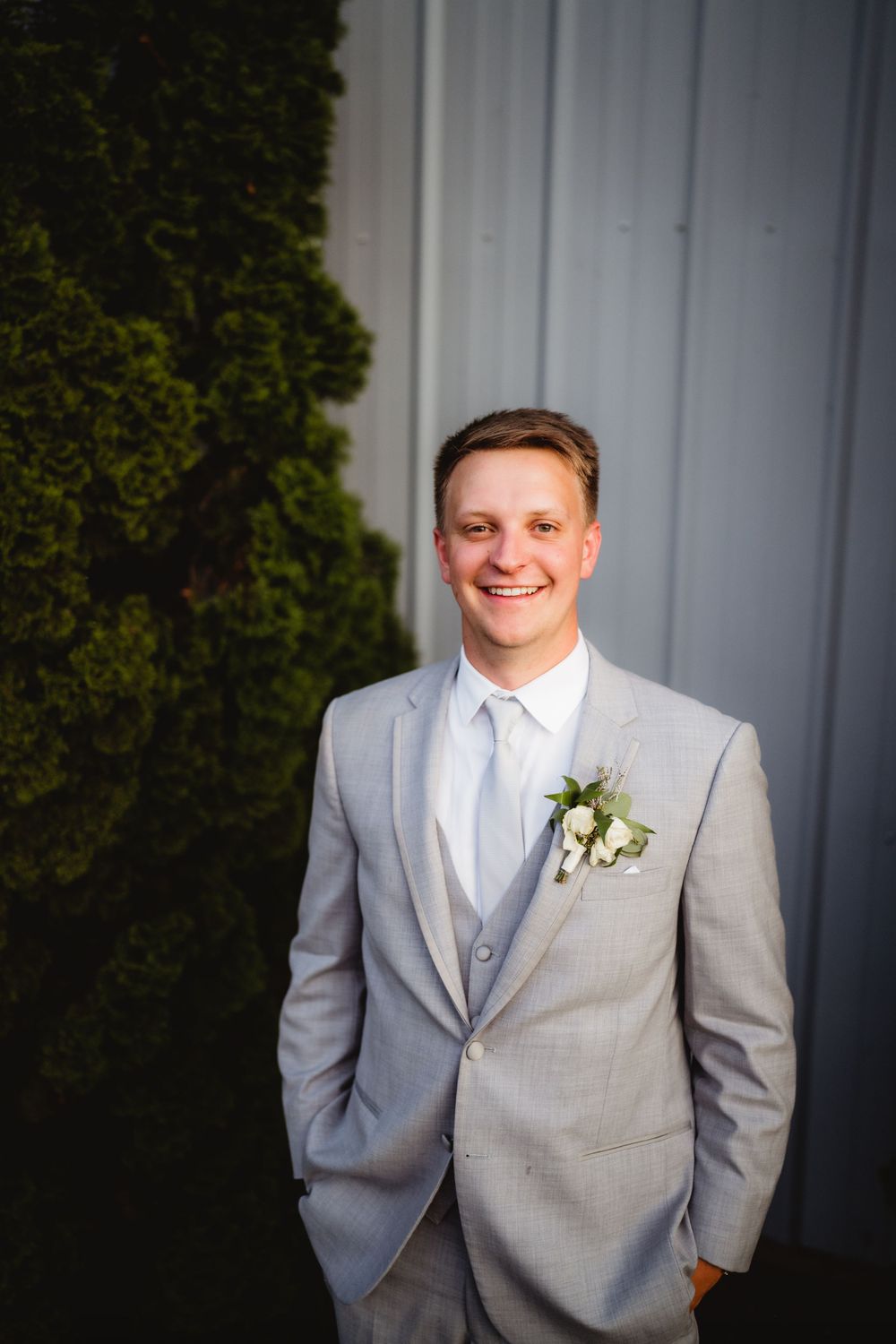 A groom in a light grey suit with a boutonniere poses against a metal wall.