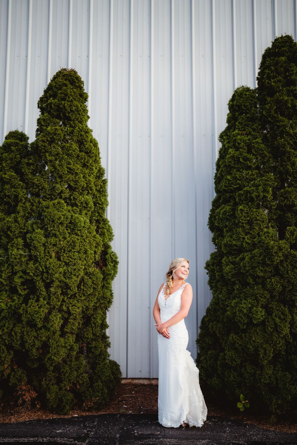 A bride in white poses between tall evergreen trees against a metal wall background.
