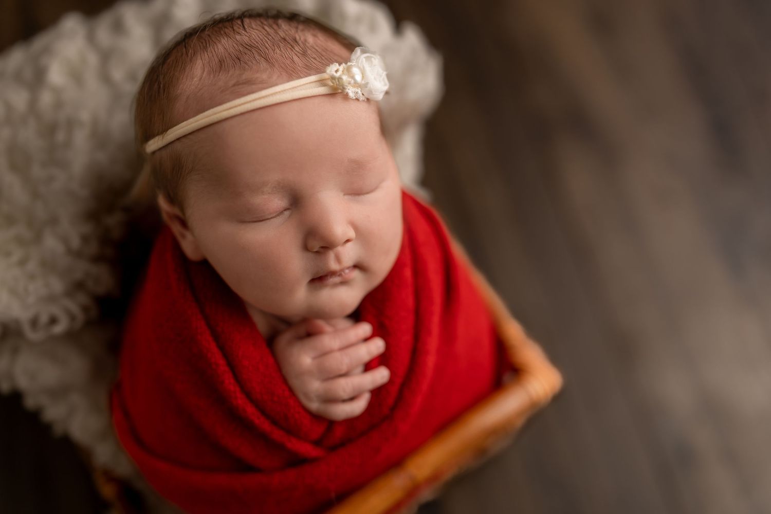 Sleeping newborn wrapped in red blanket wearing delicate headband rests in wooden bowl.
