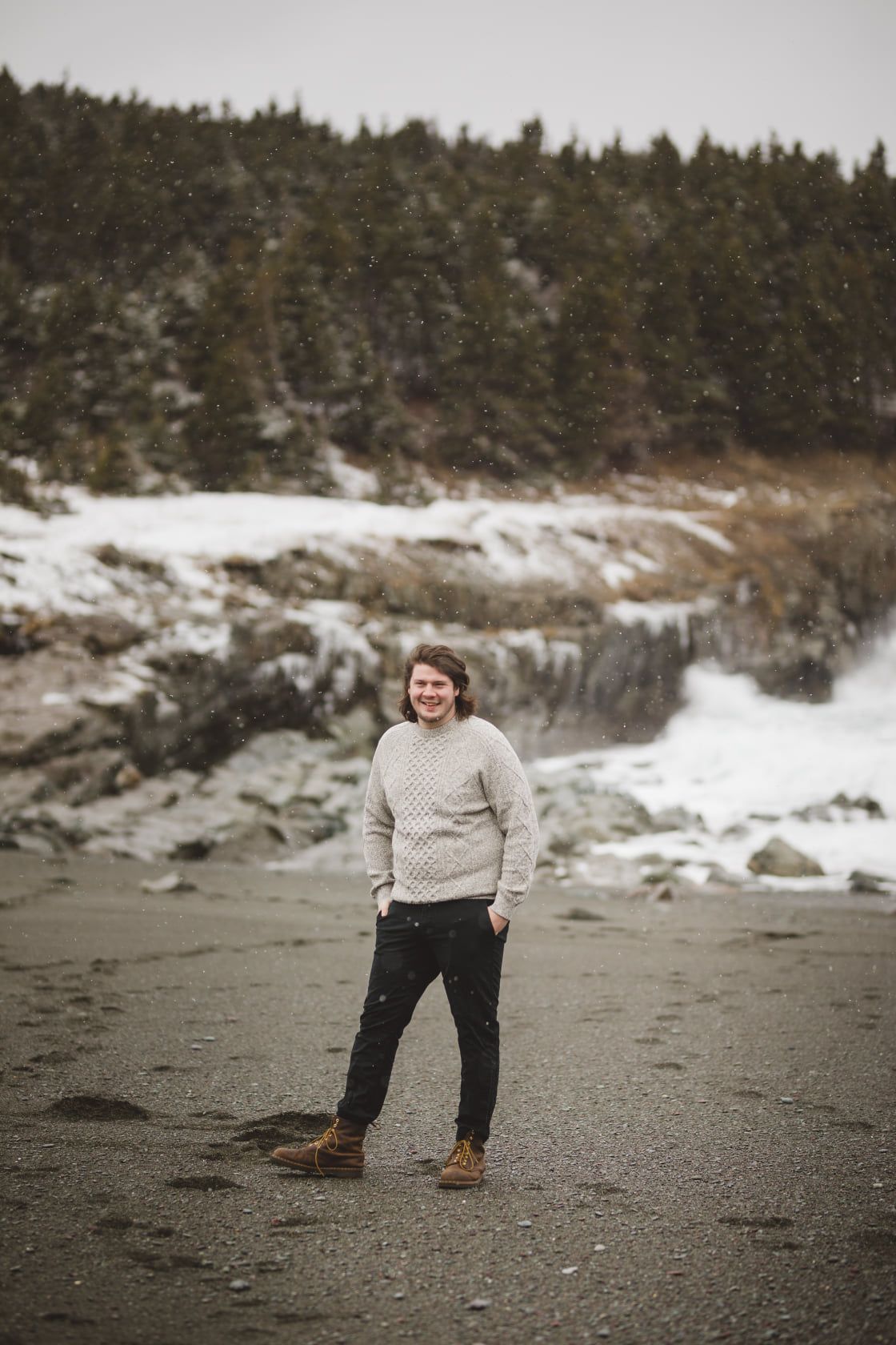 A person in a cream sweater stands on a snowy beach with evergreen forest backdrop.