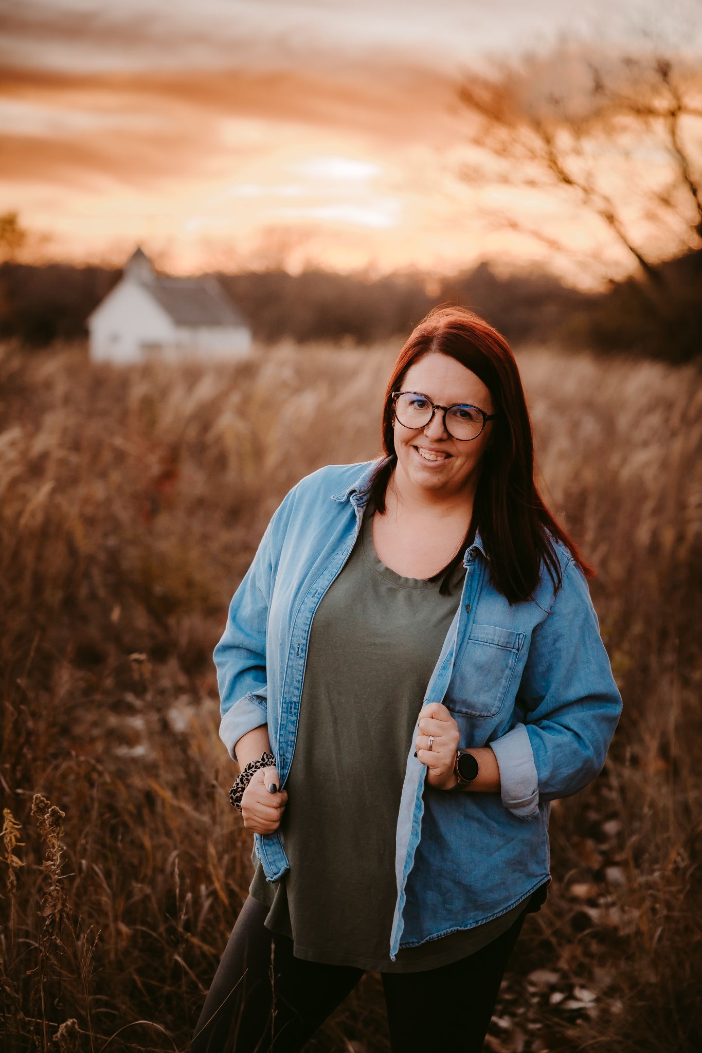 Red-haired woman in denim shirt smiling in golden field at dramatic sunset.
