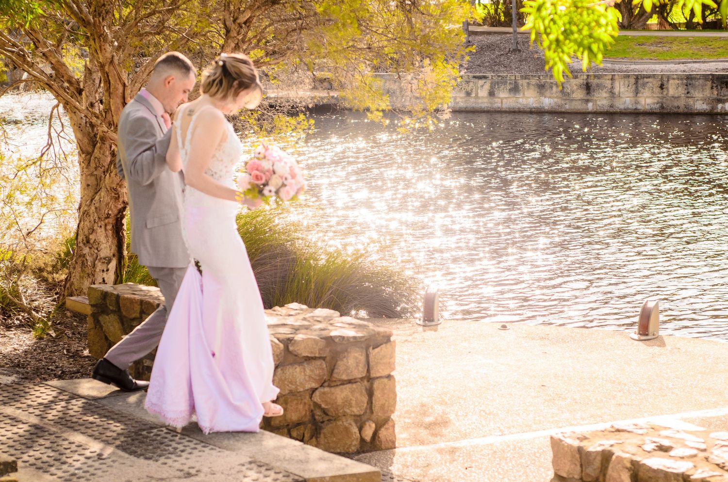 A couple stands by a waterfront stone wall in golden sunset light.