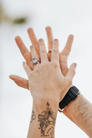 Two hands with engagement ring and tattoo overlapping against white background.