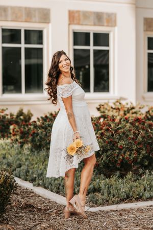 A bride in a white lace high-low dress poses outdoors with yellow sunflowers.