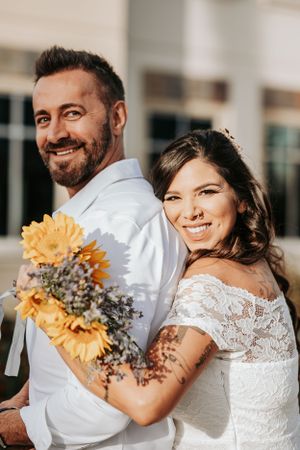 A couple poses back-to-back holding sunflowers against building architecture.