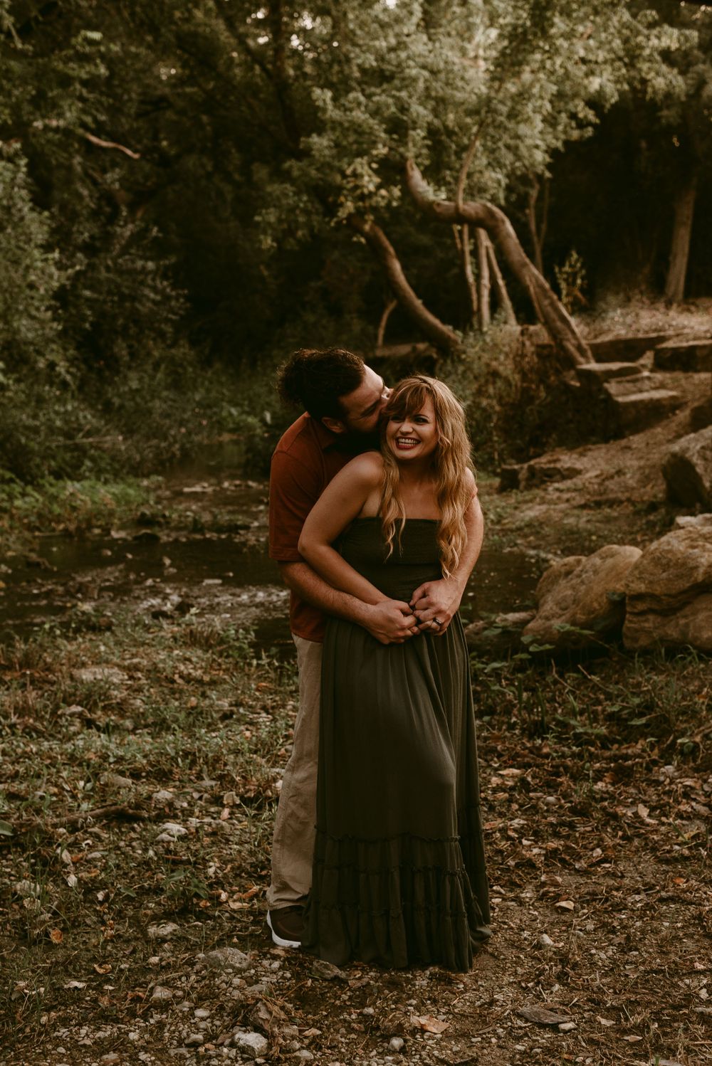 Couple embracing in a wooded area wearing coordinated rust and dark colored outfits.