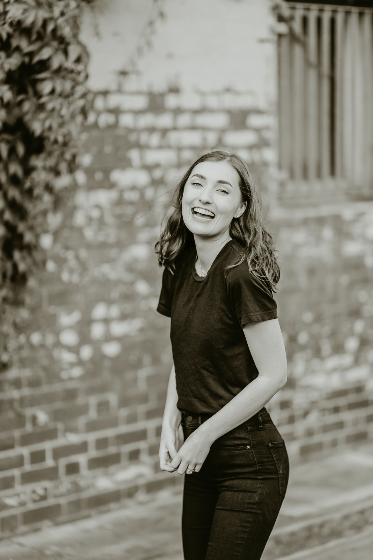 Black and white portrait of a joyful person in casual attire laughing against a brick wall background.
