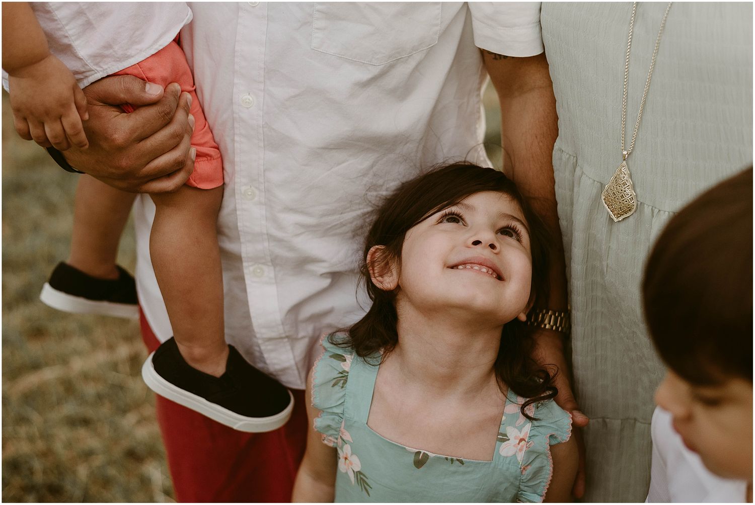 A young child in a teal dress smiles joyfully while looking up at family members during an outdoor photoshoot.