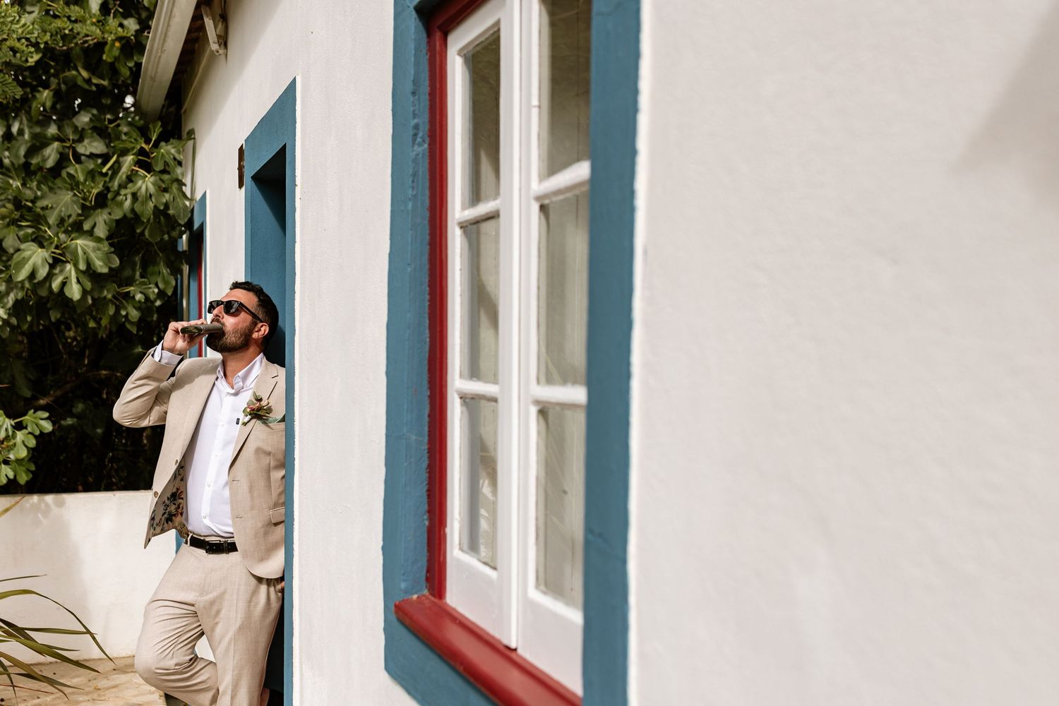 Groom in a light colored suit leans against a white wall with blue and red trim near a bright window at Quinta da Bichinha