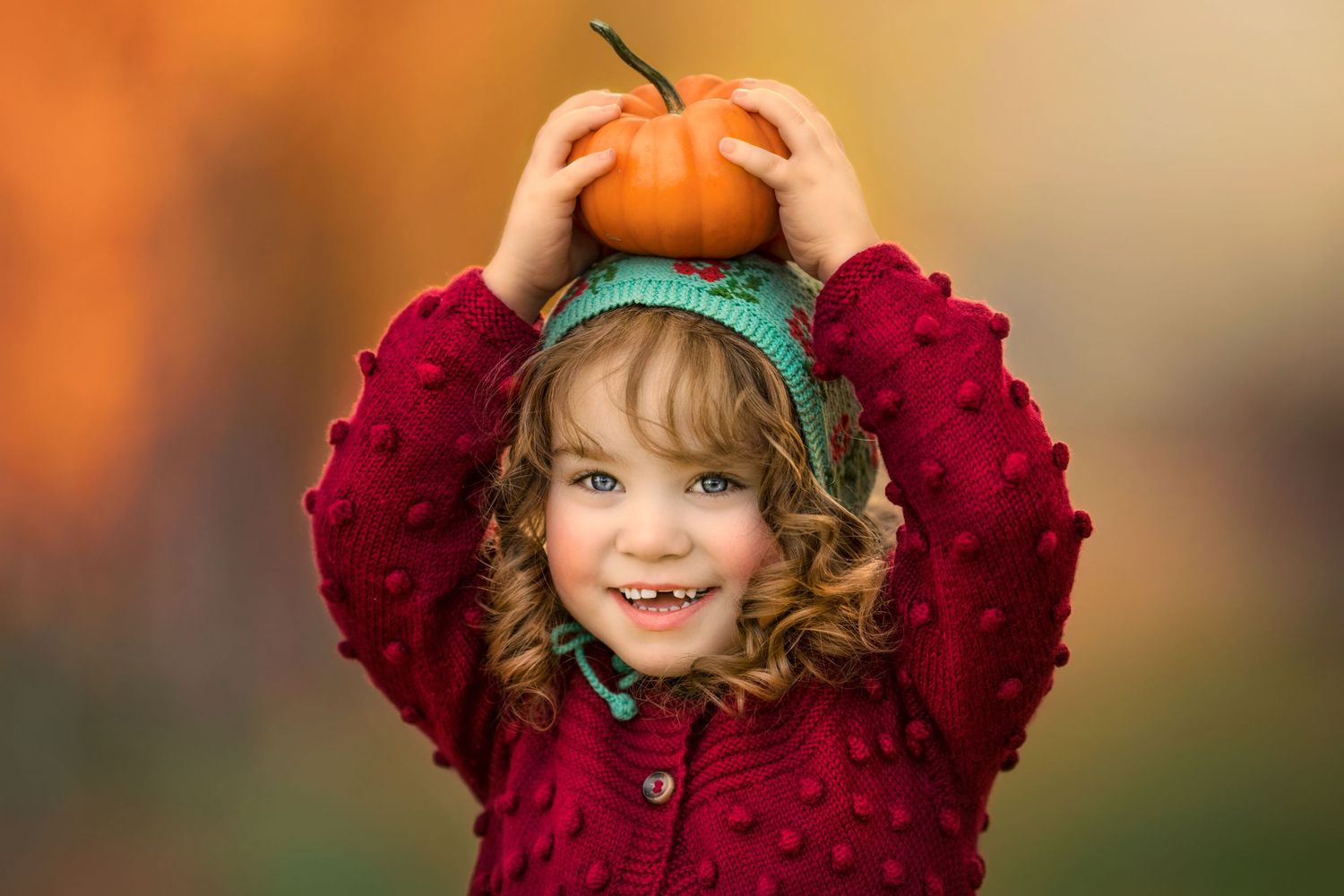 Joyful autumn portrait showcasing a child with curly hair in red sweater holding a small pumpkin on her head.