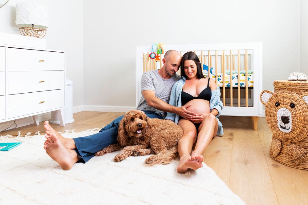 In-home maternity photoshoot with Couple relaxing on nursery floor with their fluffy golden doodle dog beside a white baby crib.