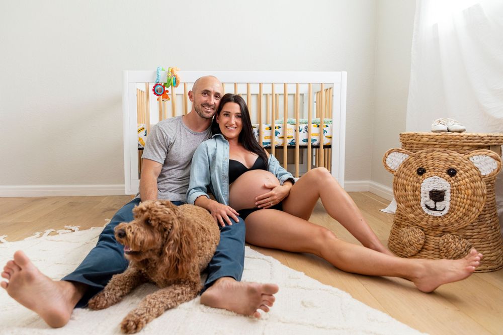 In-home maternity photoshoot with  sitting on nursery floor with golden doodle dog in front of white crib during maternity photo session.