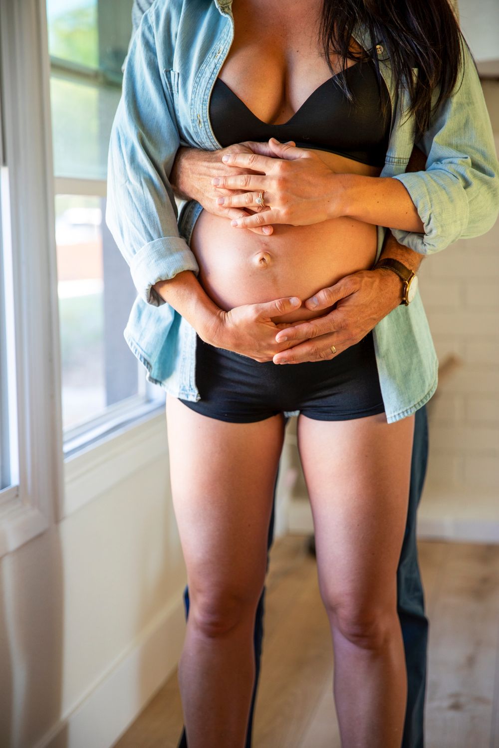 In-home maternity photoshoot with Close-up of man tenderly cradling pregnant woman's bare baby bump near sunlit window.