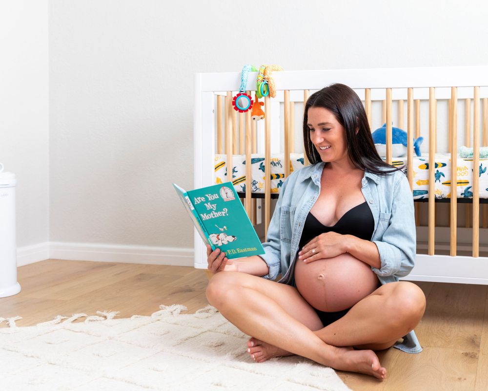 In-home maternity photoshoot with Pregnant woman sitting cross-legged on nursery floor reading a book while cradling her baby bump.