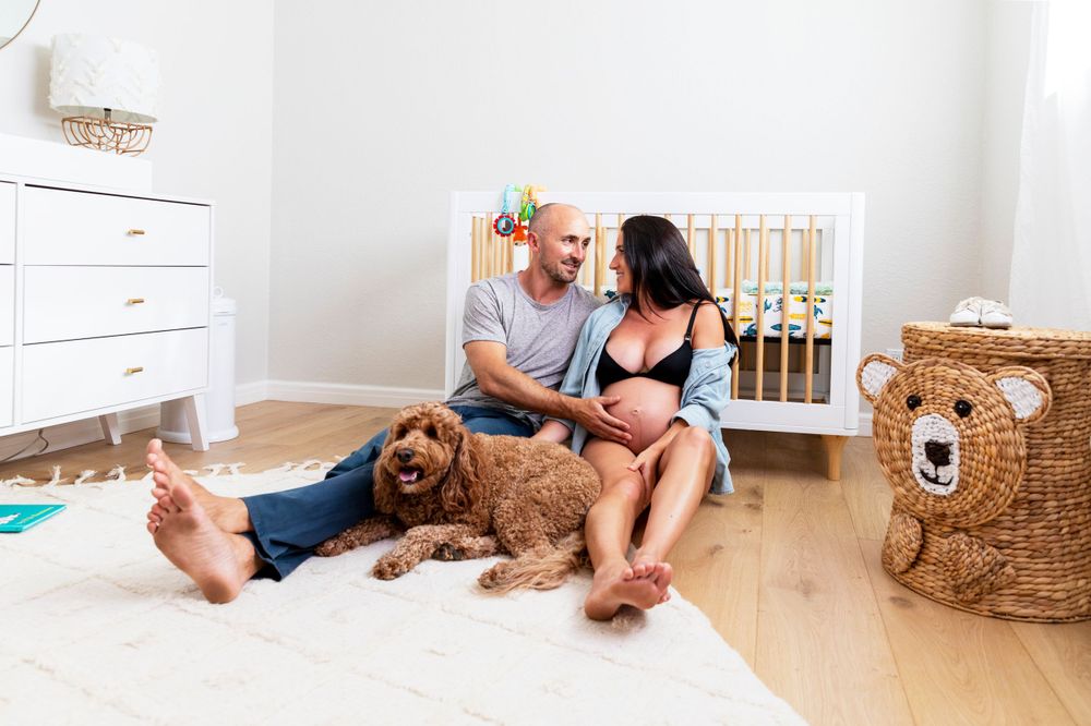 In-home maternity photoshoot with Expectant couple sitting together on nursery floor with golden doodle dog near white baby crib.