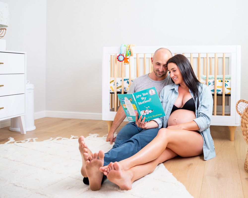 In-home maternity photoshoot with  sitting on nursery floor reading Dr. Seuss book together beside white baby crib.