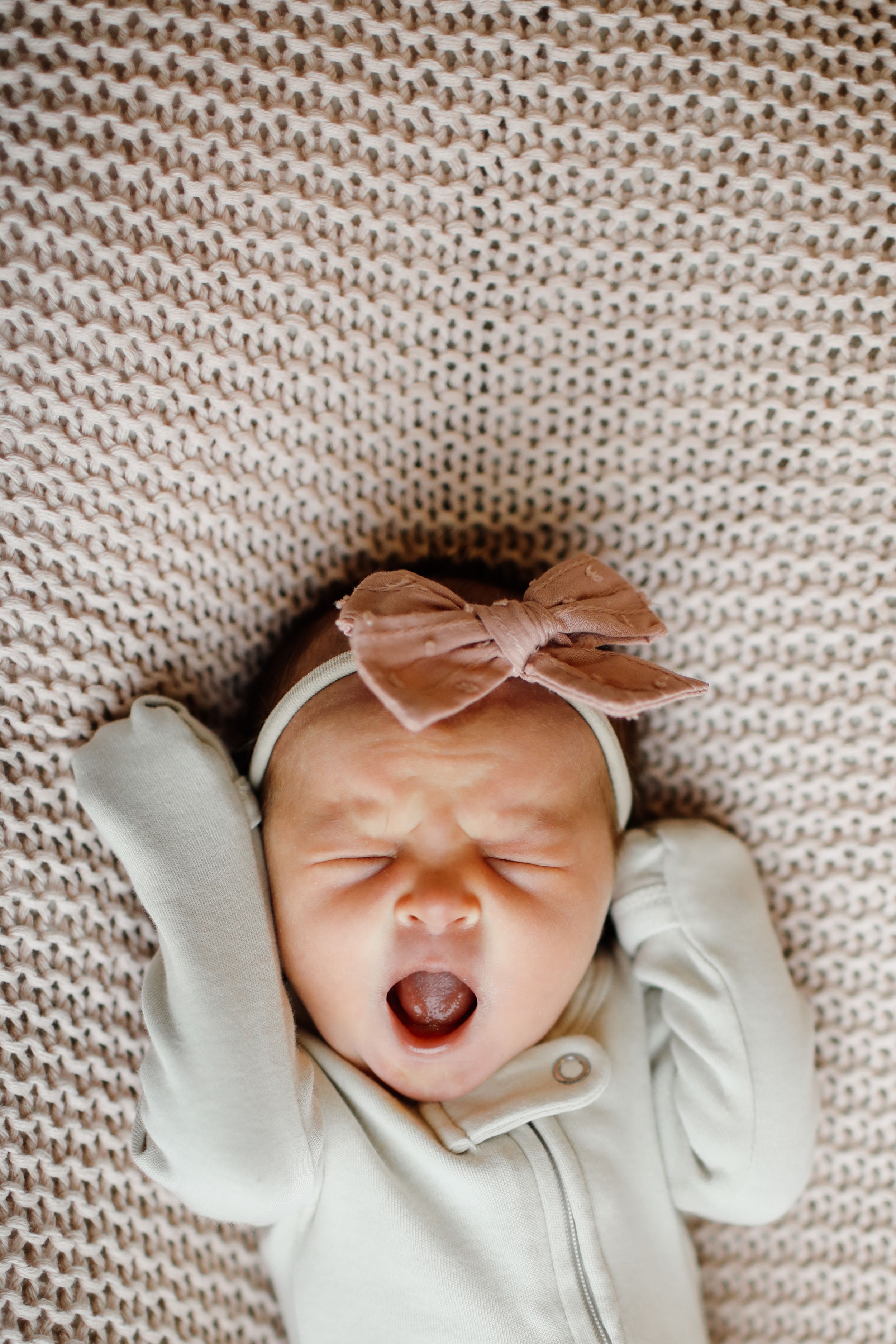 Newborn baby wearing white outfit and bow headband yawning on knit blanket.