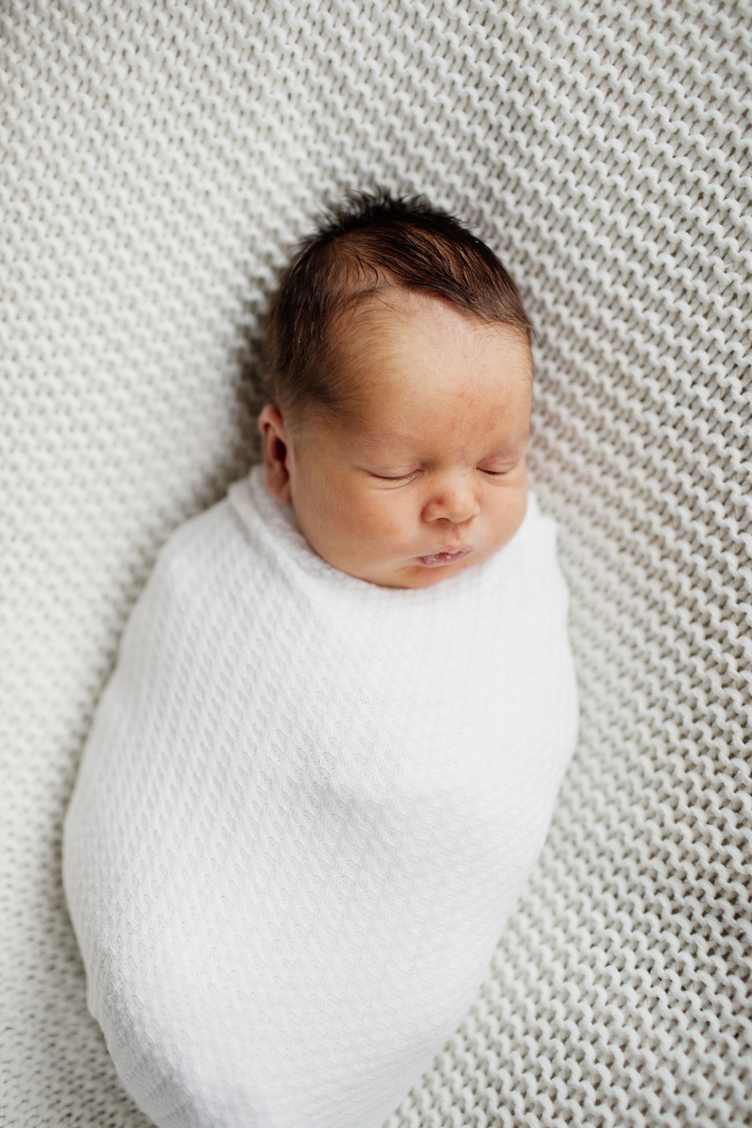 Newborn baby peacefully sleeping while swaddled in white blanket on textured gray background.