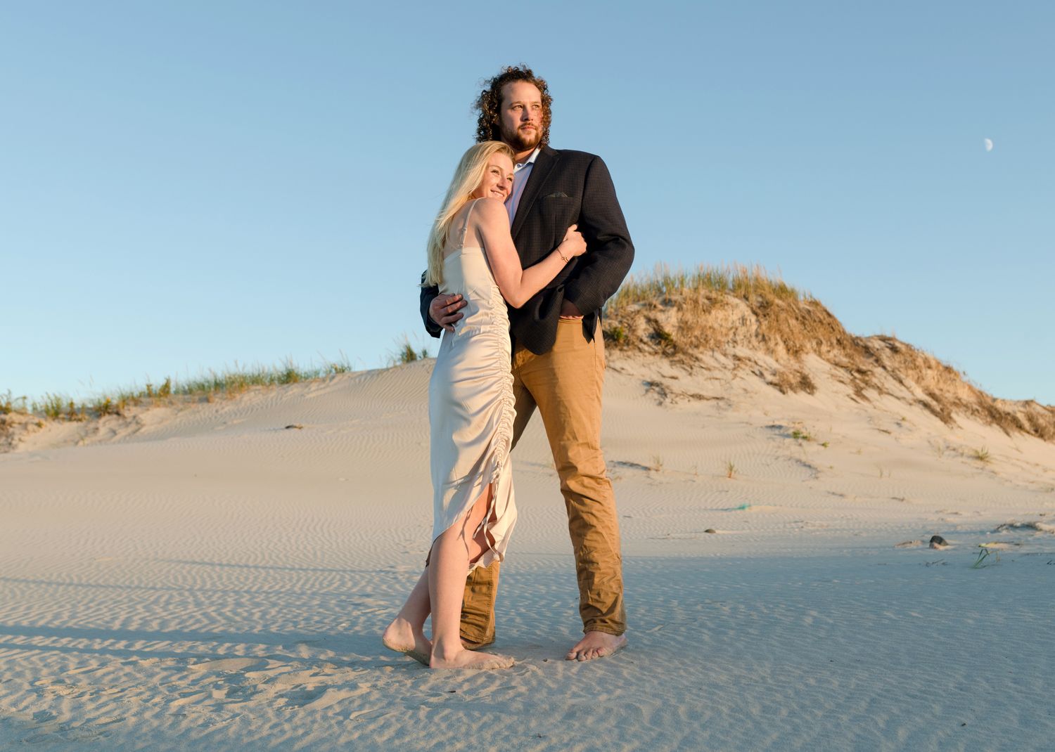 Couple embracing on sandy dunes during golden hour at the beach.