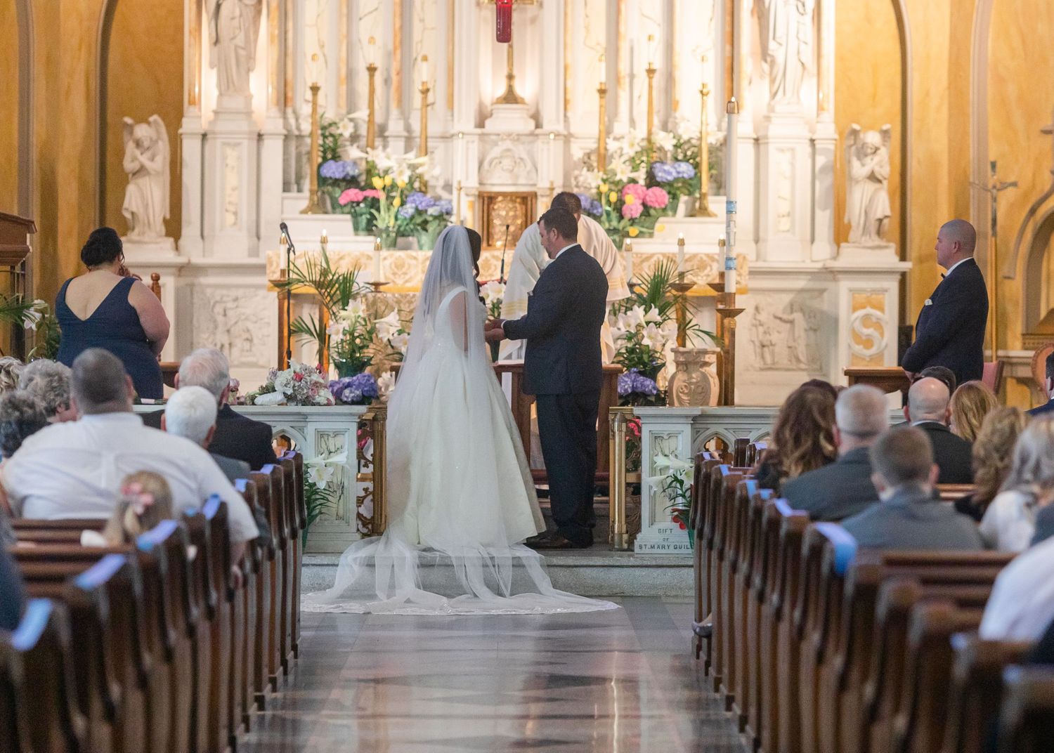 Wedding ceremony in progress at ornate Catholic church with wooden pews and decorative altar.