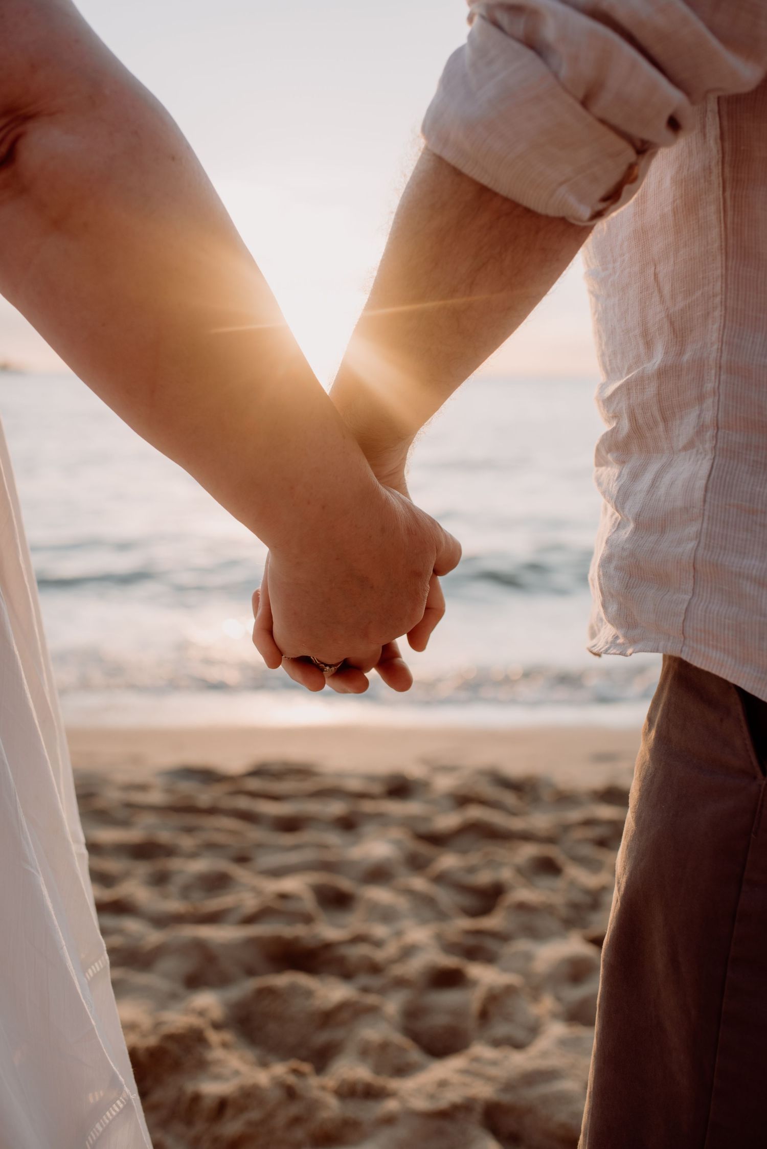 A romantic close-up of two people holding hands on a sandy beach at sunset with sunlight filtering through.