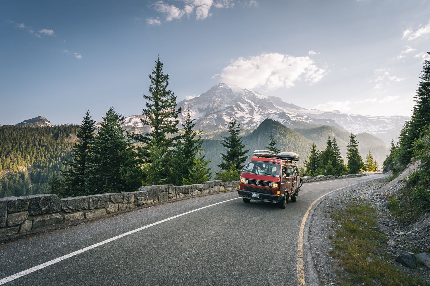 Red vintage car drives along scenic mountain road with Mount Rainier towering in background.