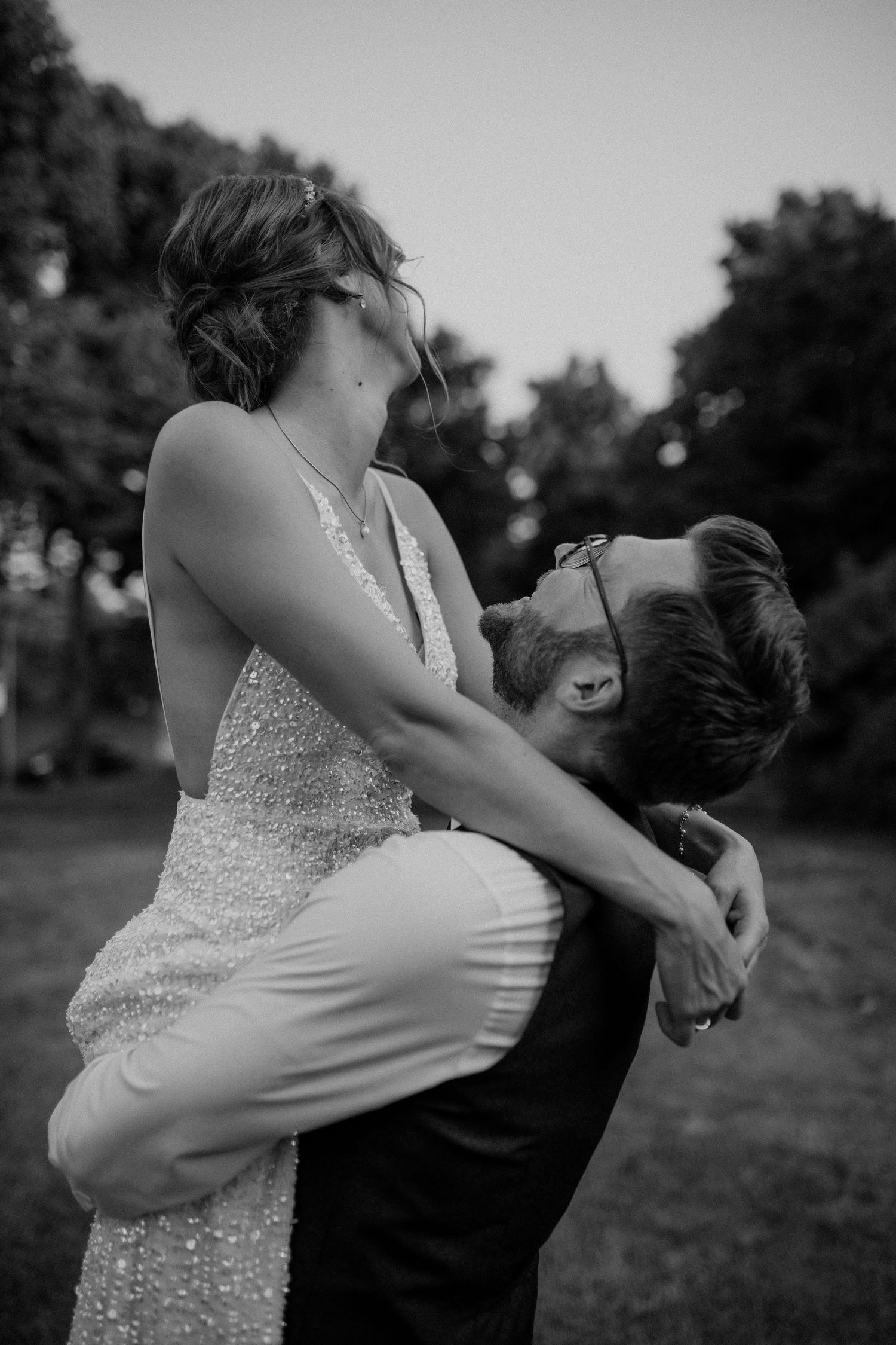 Black and white photo of groom lifting bride in sparkly wedding dress outdoors.