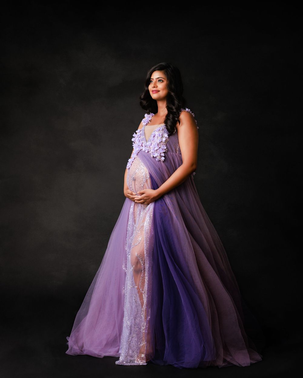 Dramatic maternity photo in purple tulle gown with lace details against dark background.