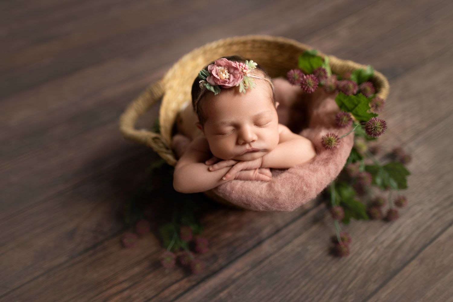 Sleeping newborn with floral headband resting in rustic bowl with greenery on wooden surface.