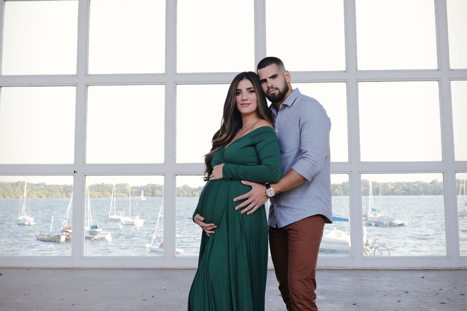 Couple posing for maternity photo, woman in green gown by waterfront windows.