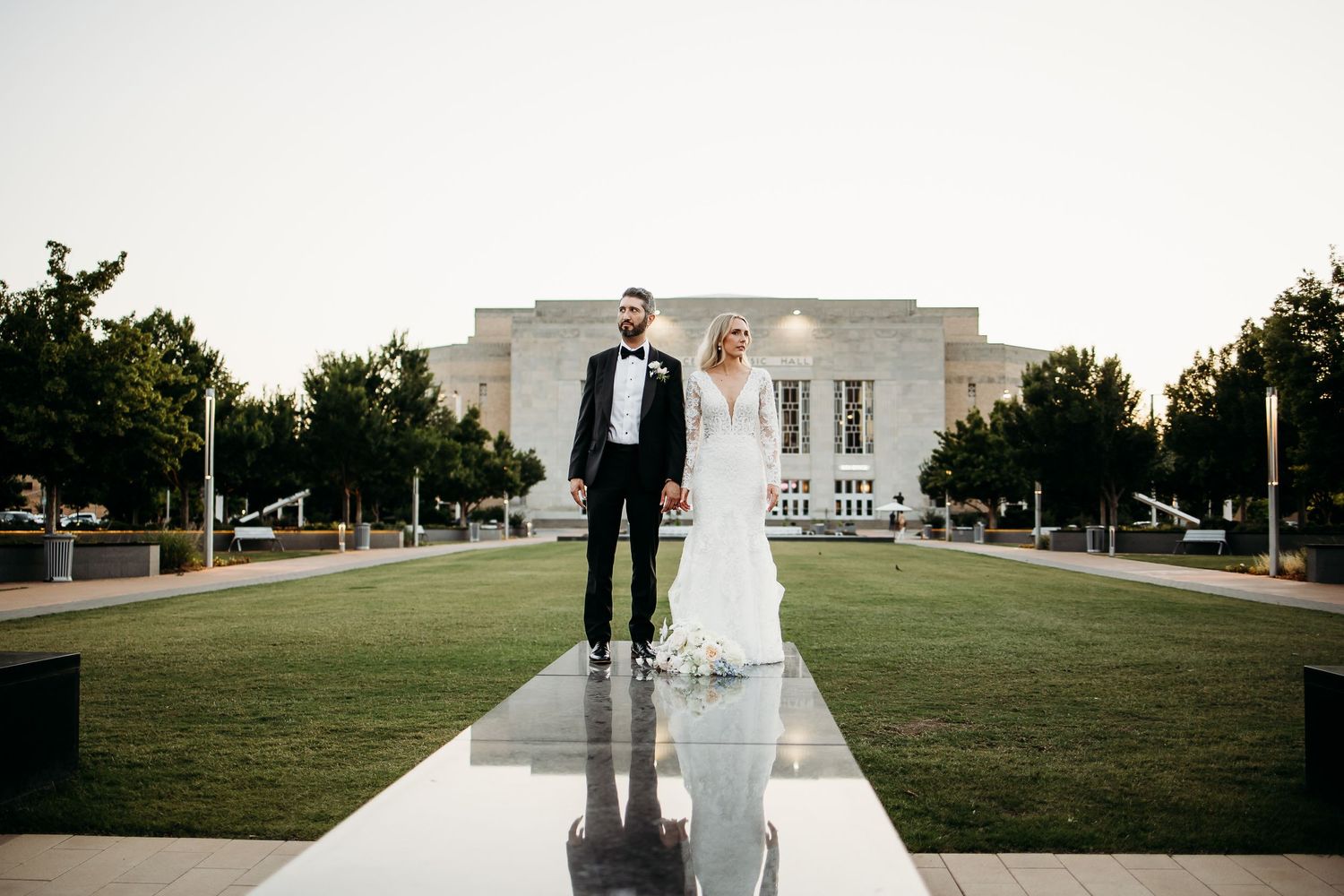 A couple in wedding attire standing on a reflective surface in front of a grand building at dusk.