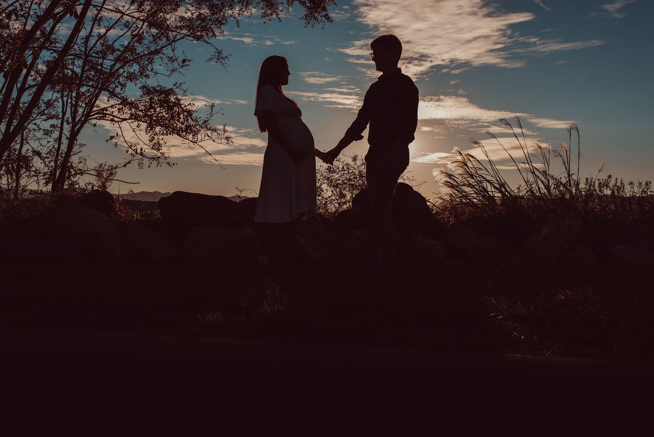 Outline of man and pregnant woman holding hands with sunset behind them during maternity photoshoot.