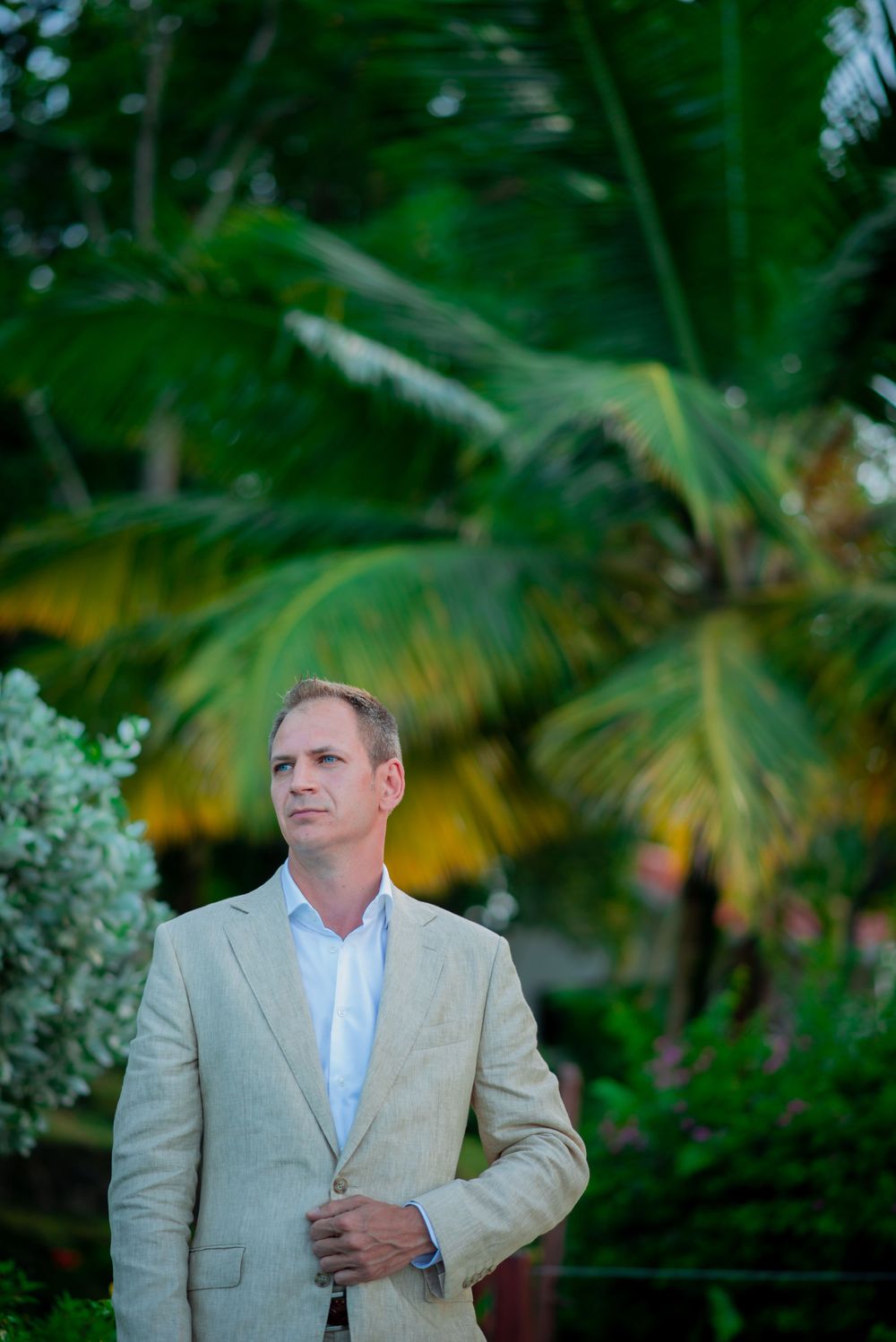Man in beige suit poses thoughtfully in tropical garden setting with palm trees.