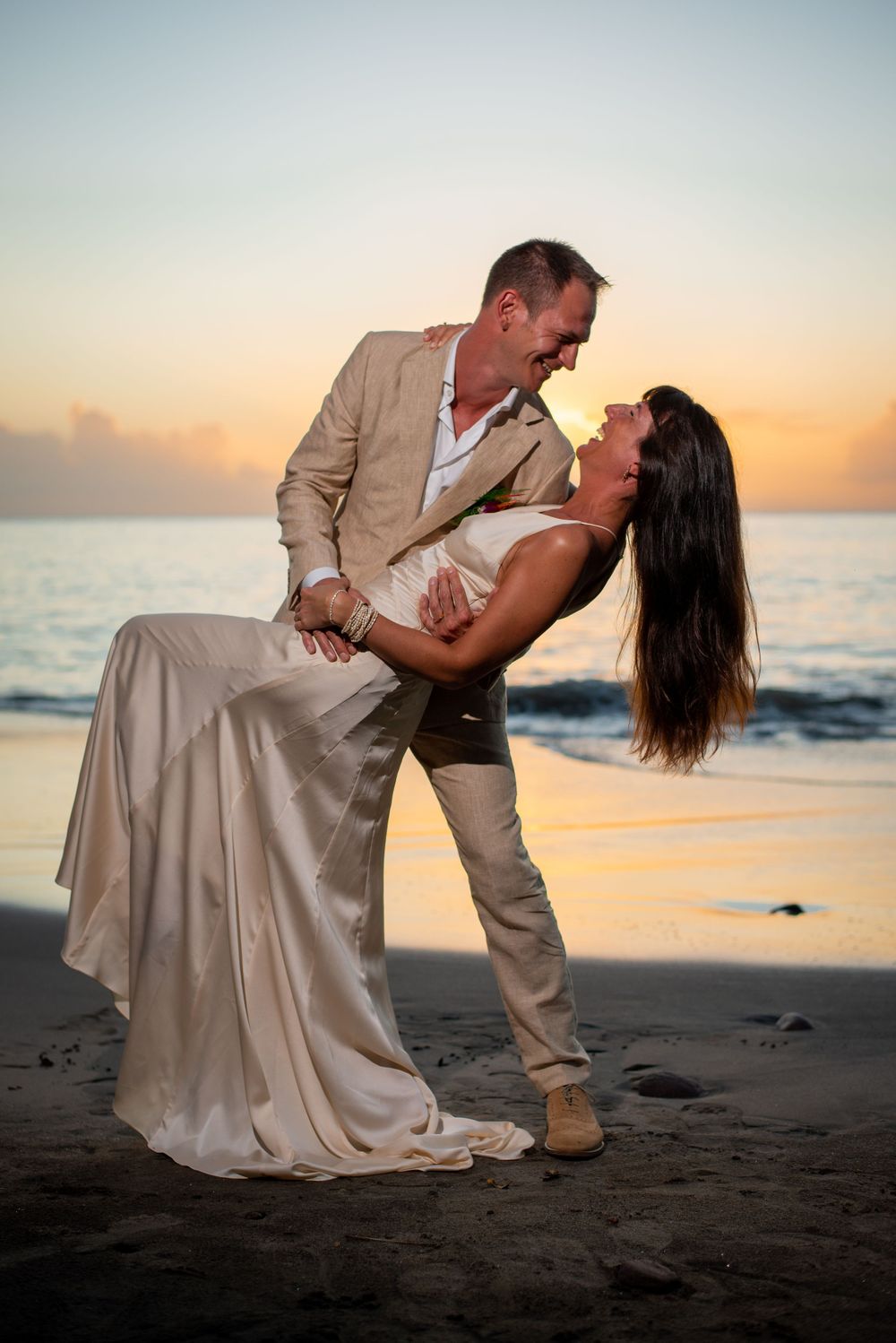 Romantic beach photoshoot at sunset with couple in formal attire on dark sand.