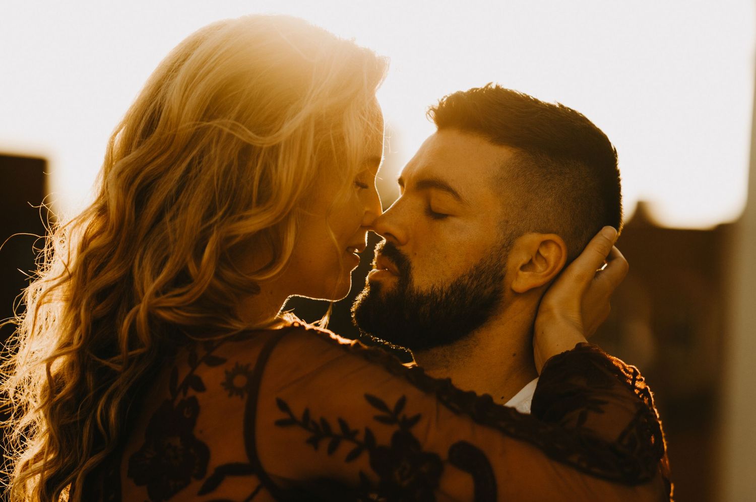 Couple sharing an intimate kiss bathed in warm golden backlight, woman with floral lace tattoo.