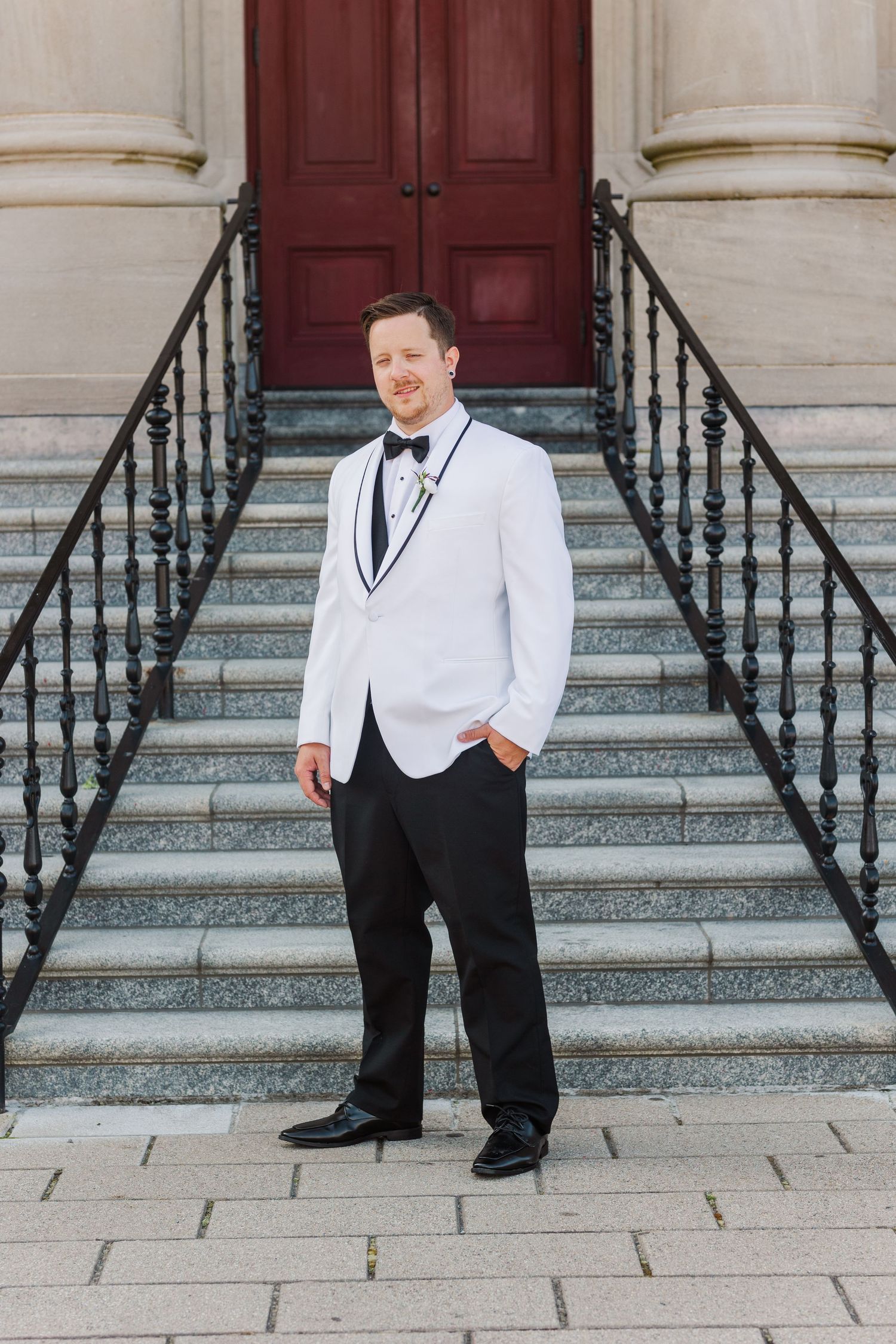 Wedding party member in white tuxedo and black pants poses on stone steps.